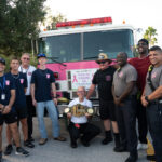 A diverse group of firefighters and supporters pose in front of a firetruck decorated with a "Pink on Parade" banner One person is holding a gold championship style belt The group looks happy and united, participating in an event to raise awareness for breast cancer