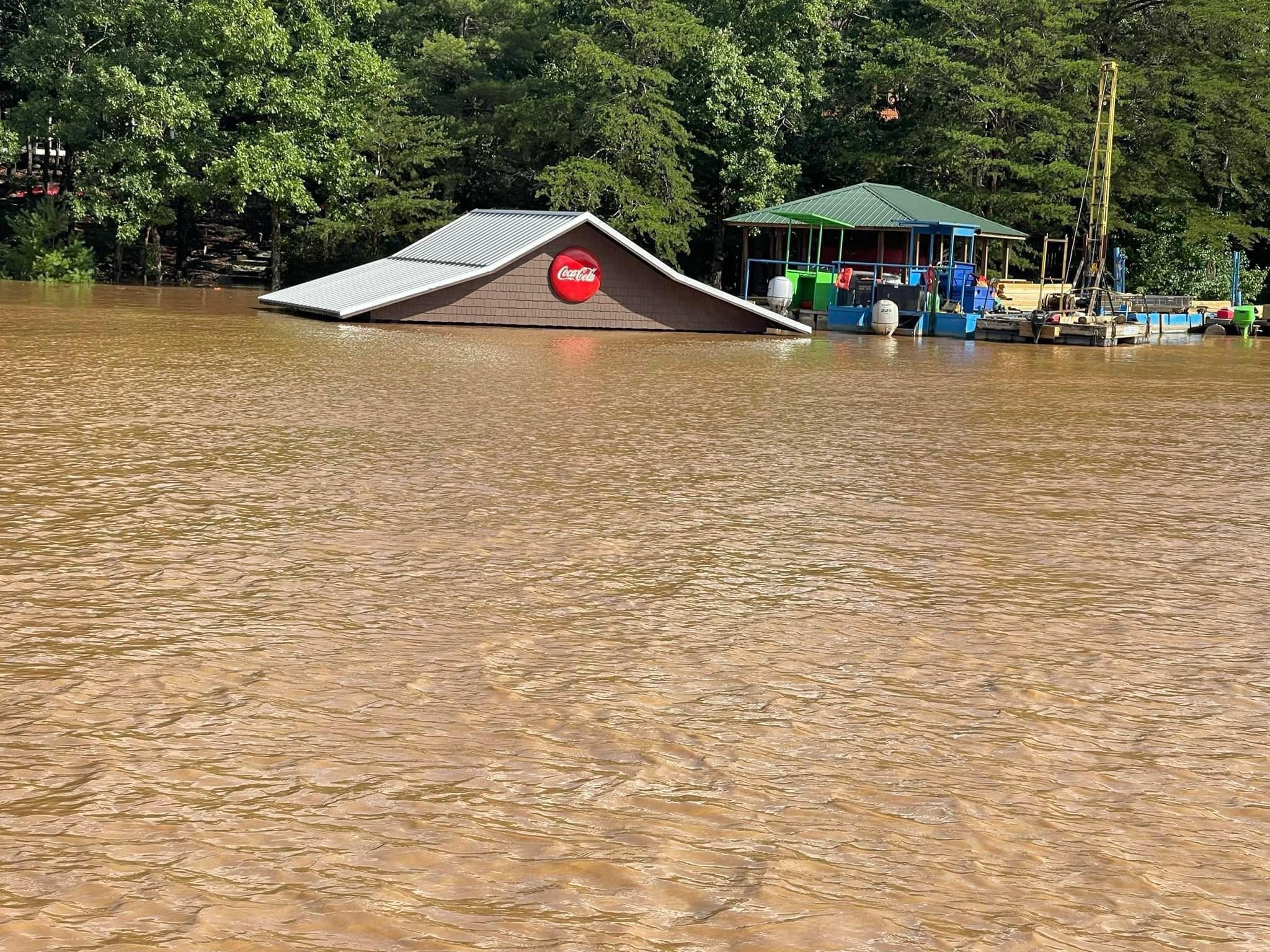 A heavily flooded area where only the roof of a building with a Coca Cola logo is visible above the water Nearby, there is another partially submerged structure with colorful equipment, surrounded by trees