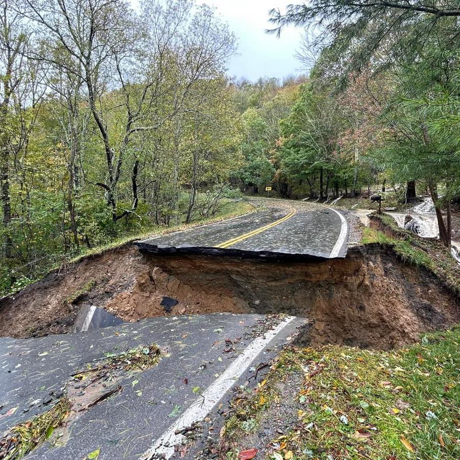 A road through a forested area has collapsed, creating a large sinkhole that spans its entire width Trees with green and yellowing leaves are visible on either side of the road The sky is overcast, and debris is scattered near the edge of the sinkhole