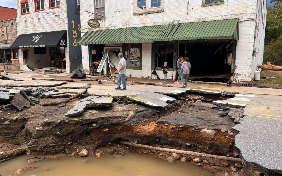 A damaged street with a large section of the road collapsed, creating a hole filled with muddy water Three people stand near the edge of the collapse, observing the damage Buildings in the background show signs of wear, with debris scattered around