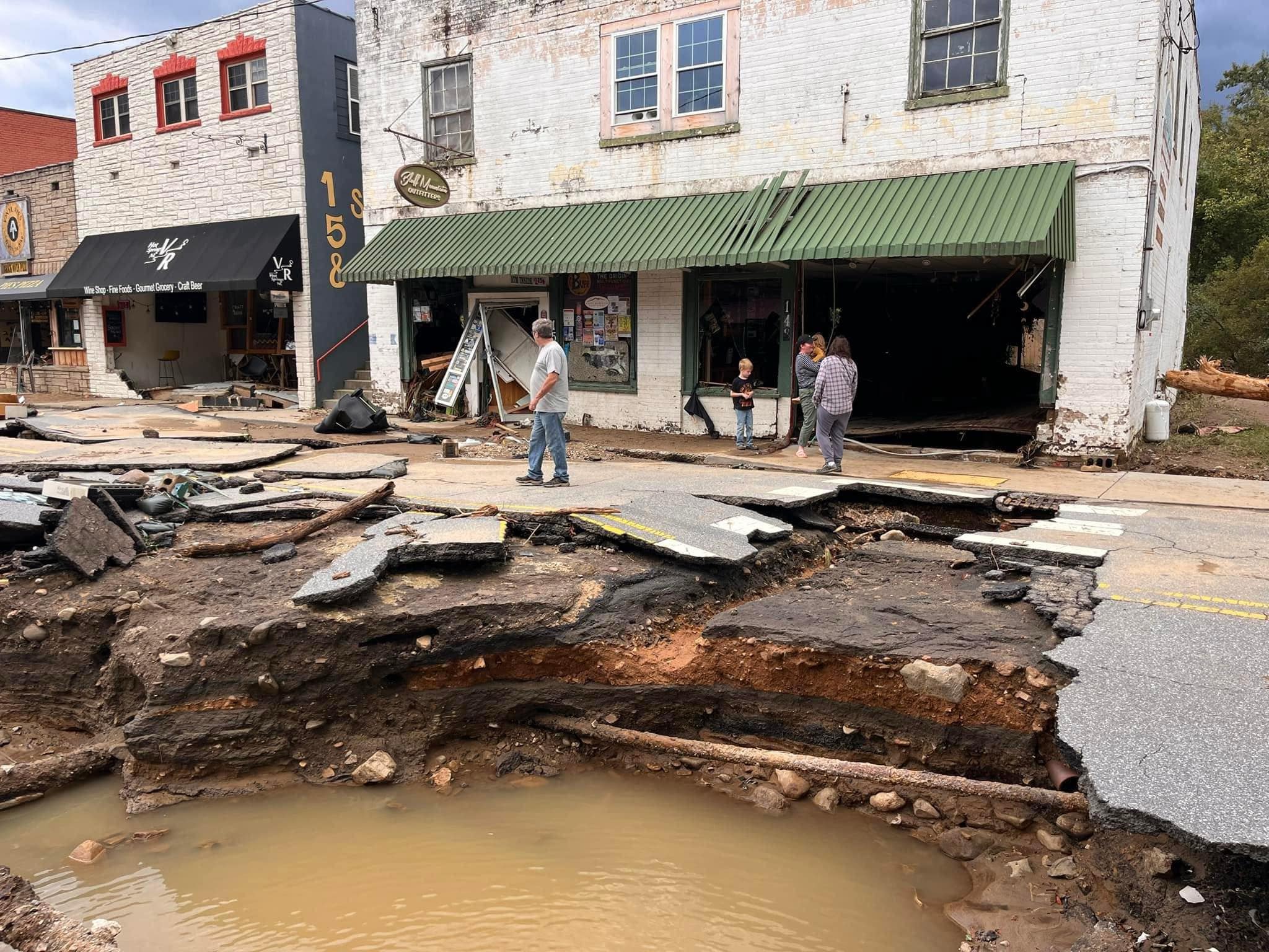 A damaged street with a large section of the road collapsed, creating a hole filled with muddy water Three people stand near the edge of the collapse, observing the damage Buildings in the background show signs of wear, with debris scattered around