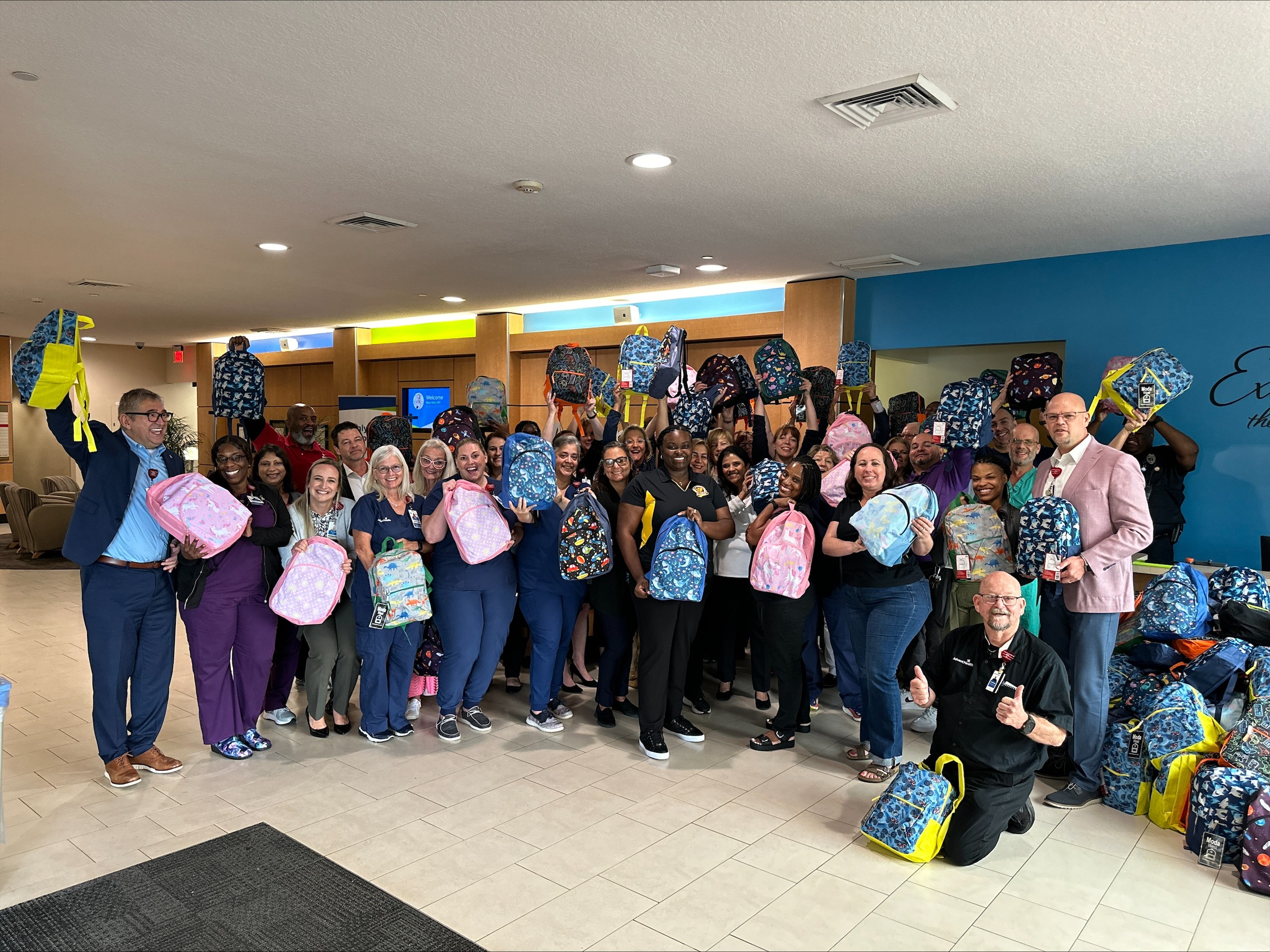 A large group of adults stand together indoors, smiling and holding up colorful backpacks The room has white walls and tiled floors, with a blue wall in the background displaying part of a logo Most people are dressed in casual or business casual attire