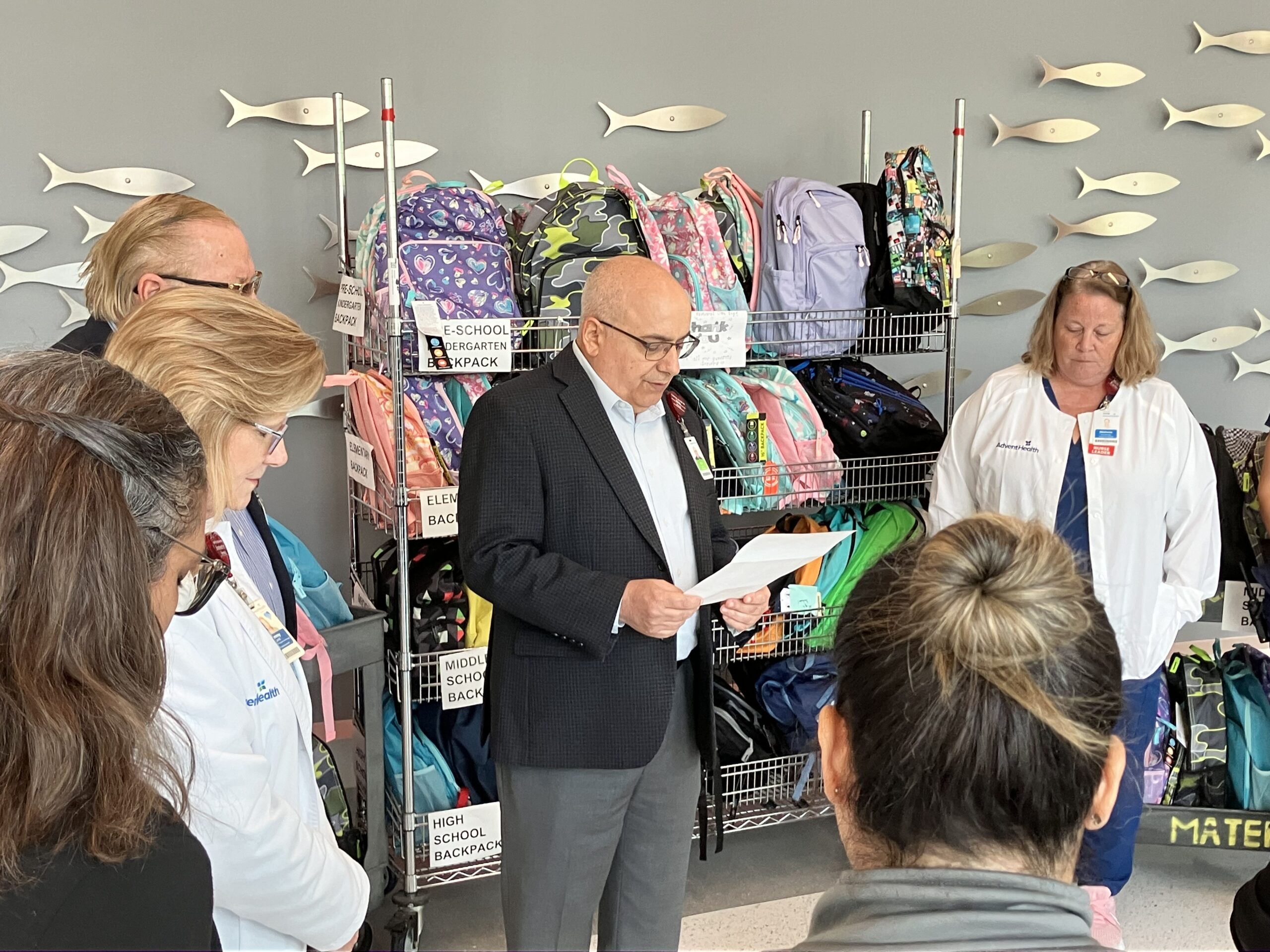 A group of people stand in front of shelves filled with colorful backpacks An older man in a suit reads from a piece of paper, with several onlookers around him, some wearing white coats The environment suggests an organized event or gathering