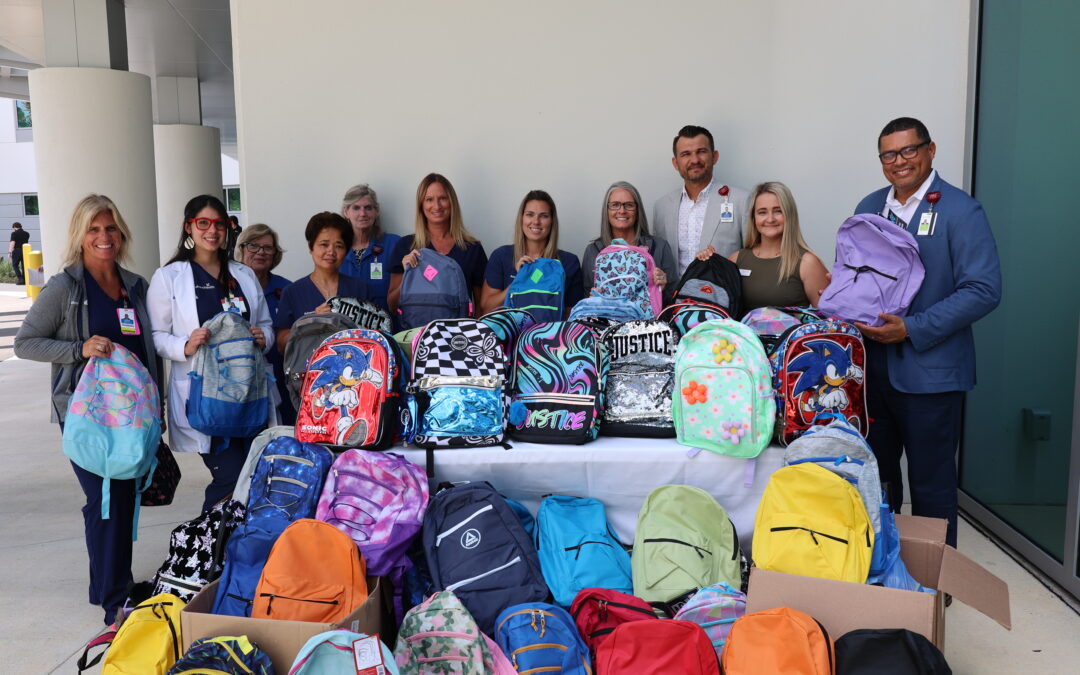 A group of people are standing behind a table filled with colorful backpacks They are dressed in professional and casual attire, some wearing name badges The background features a modern building with white columns There are boxes with additional backpacks in front