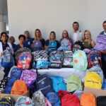 A group of people are standing behind a table filled with colorful backpacks They are dressed in professional and casual attire, some wearing name badges The background features a modern building with white columns There are boxes with additional backpacks in front