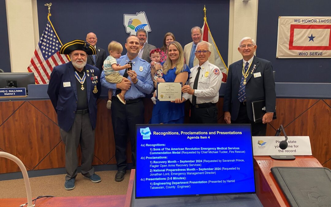 A group of people stands together in a meeting room, several of whom are in uniforms A woman in blue is holding a certificate, smiling broadly, surrounded by others who are also smiling Behind them is a screen displaying agenda items for recognitions and presentations