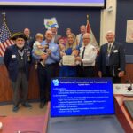 A group of people stands together in a meeting room, several of whom are in uniforms A woman in blue is holding a certificate, smiling broadly, surrounded by others who are also smiling Behind them is a screen displaying agenda items for recognitions and presentations