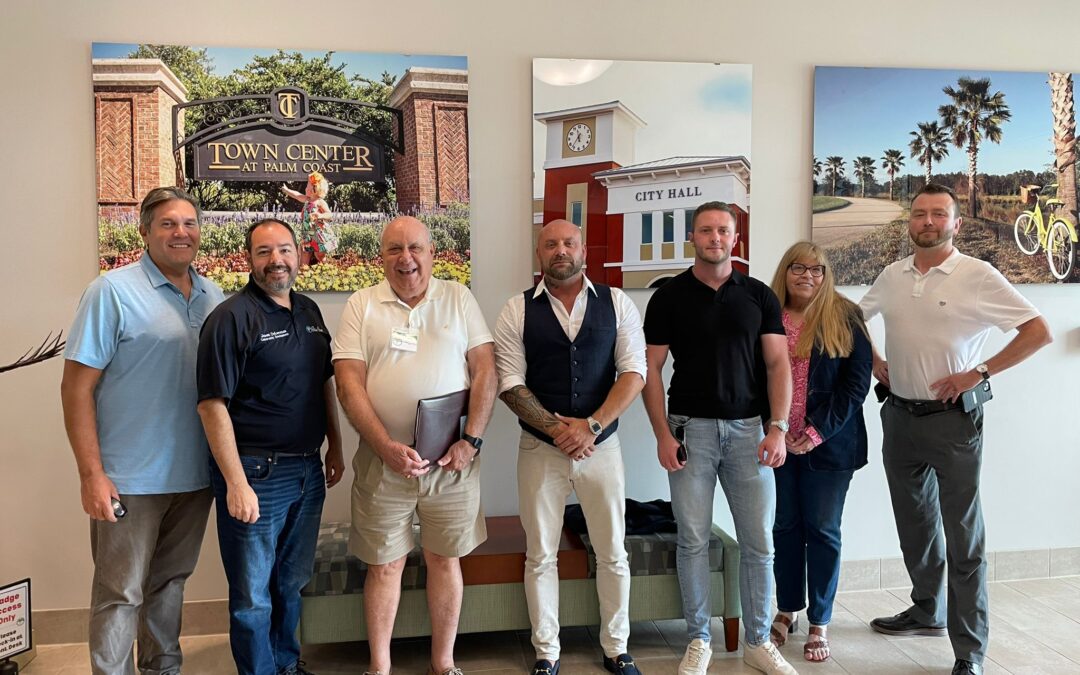 A group of seven people stands indoors posing for a photo Three large framed images of Town Center, City Hall, and a park with palm trees and a bicycle are mounted on the wall behind them They are casually dressed and smiling at the camera