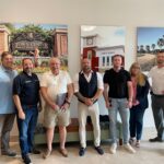 A group of seven people stands indoors posing for a photo Three large framed images of Town Center, City Hall, and a park with palm trees and a bicycle are mounted on the wall behind them They are casually dressed and smiling at the camera