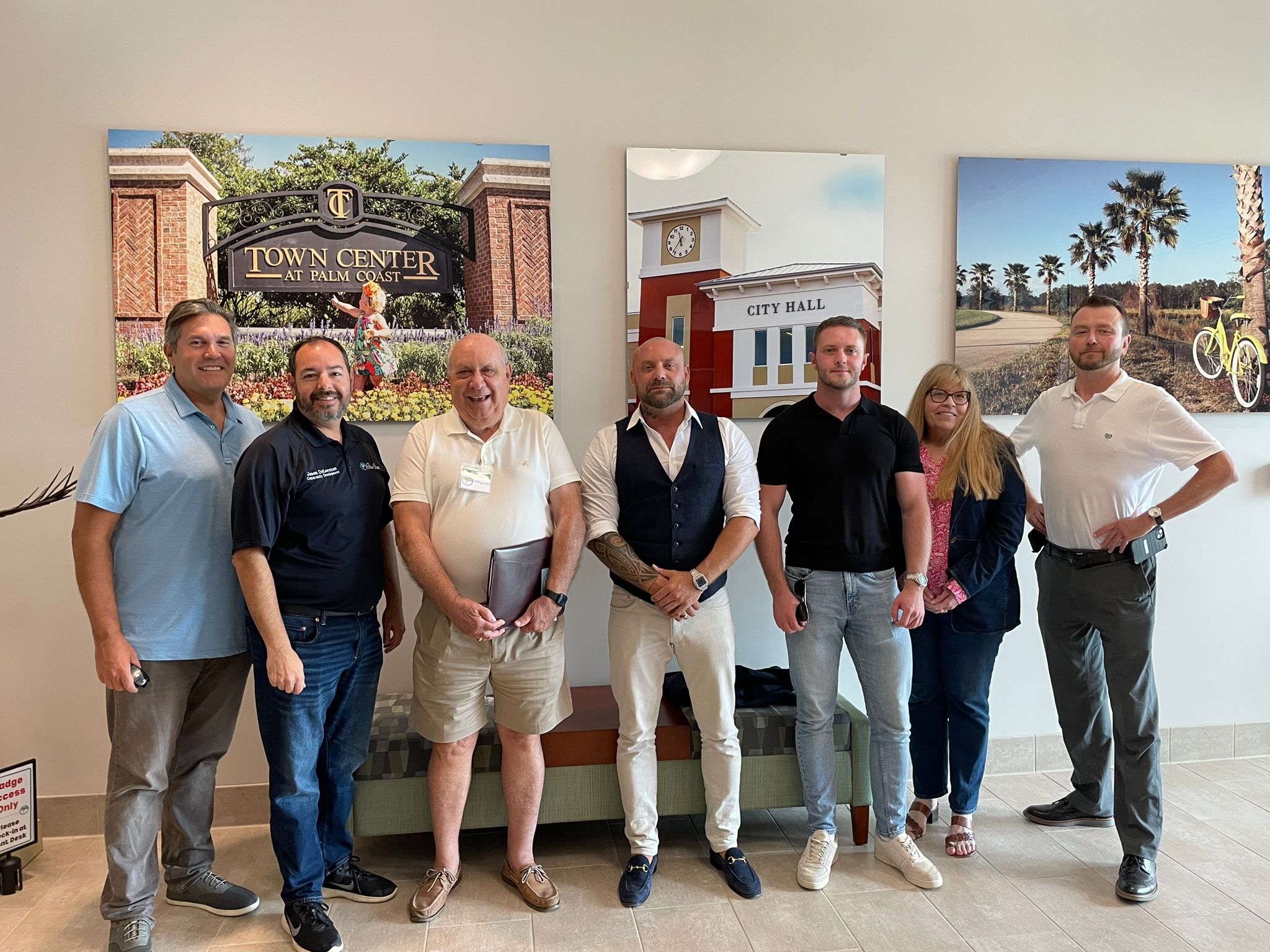 A group of seven people stands indoors posing for a photo Three large framed images of Town Center, City Hall, and a park with palm trees and a bicycle are mounted on the wall behind them They are casually dressed and smiling at the camera