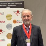 An older man with a beard and gray hair stands in front of a banner for the Florida Authors and Publishers Association President's Book Awards He is wearing a dark suit, a striped tie, and two medals around his neck