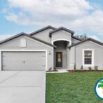 A modern, single story gray house with a well kept lawn and driveway leading to a two car garage The front door features a small, covered porch There is a logo in the bottom right corner with a palm tree and blue waves