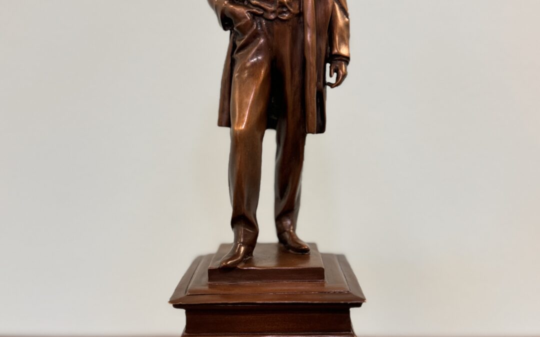 A bronze statue of a man dressed in th century attire, including a long coat and vest, stands on a rectangular wooden pedestal The man's hand is partially in his pocket, and he gazes forward with a solemn expression The background is plain and light colored