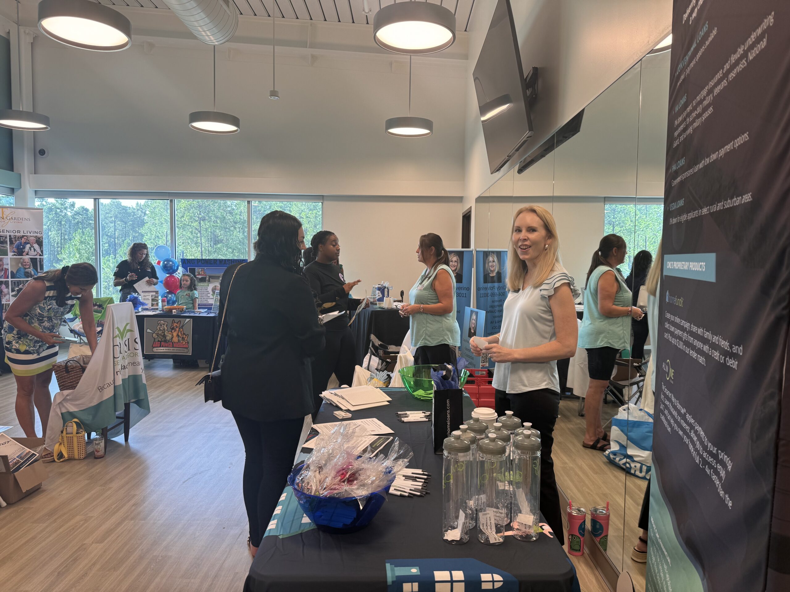 A group of people interacting at an indoor event with various booths set up A woman in a light blue blouse stands smiling at a table displaying informational materials and items Other attendees are engaged at different booths in the background