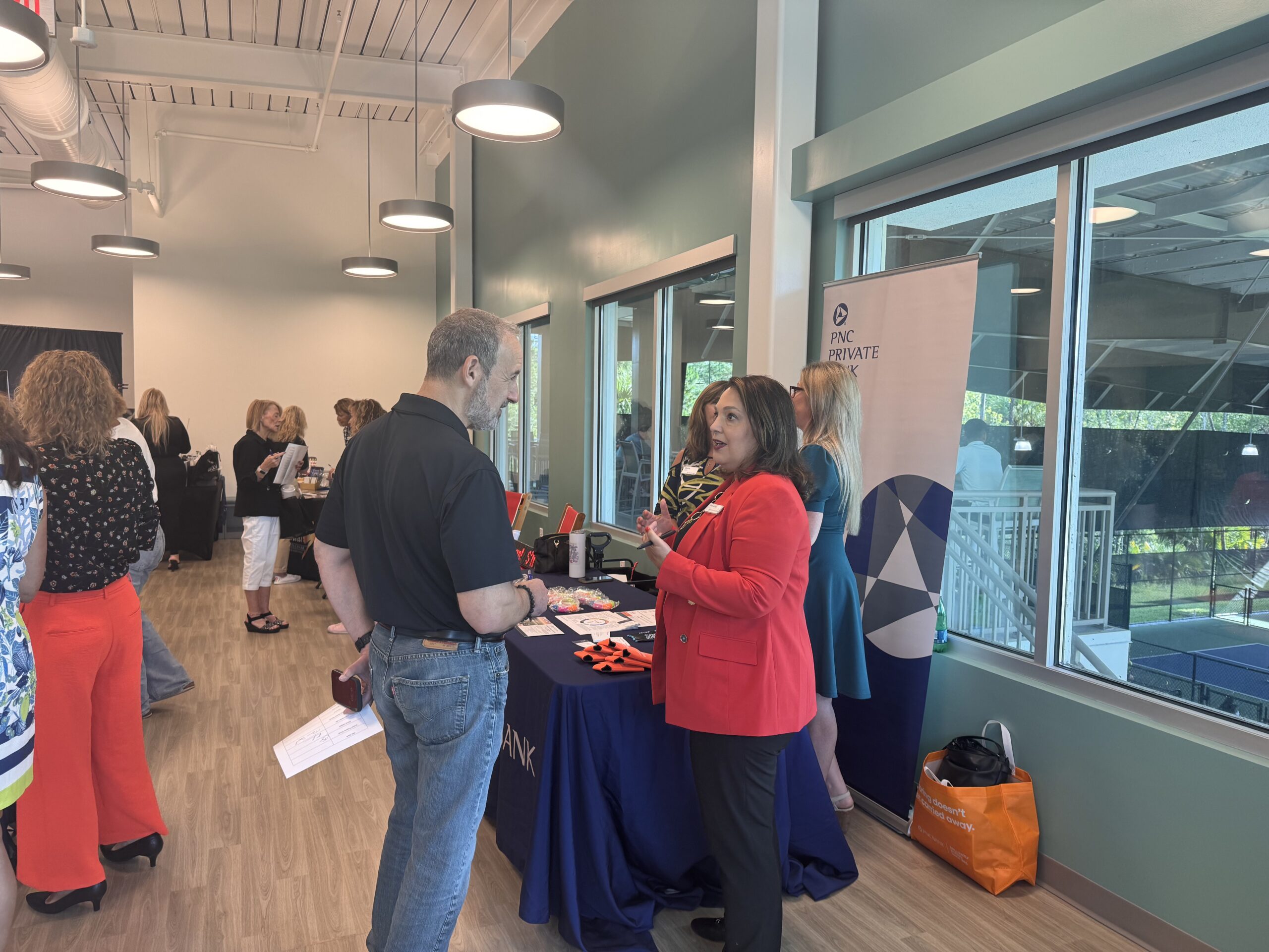 People are interacting at an indoor event with various booths A woman in a red blazer is speaking to a man in a black shirt at one booth, which has a PNC Private Bank banner Attendees are browsing and networking in a well lit room with wooden floors and large windows