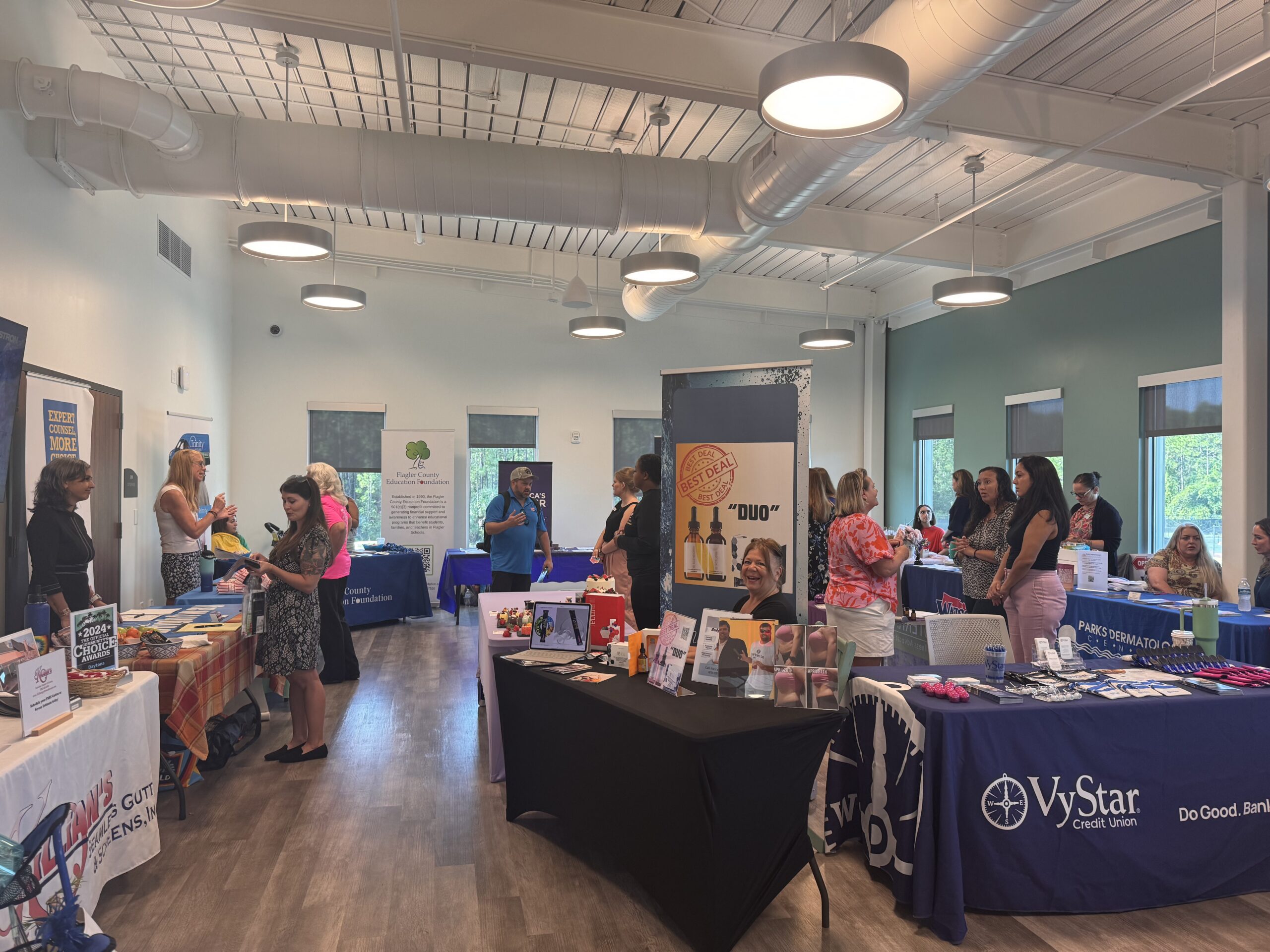 A spacious room with booths set up along the walls and people interacting at each booth Various informational materials and products are displayed, with banners and posters visible behind each booth Natural light streams in through large windows, creating a bright atmosphere