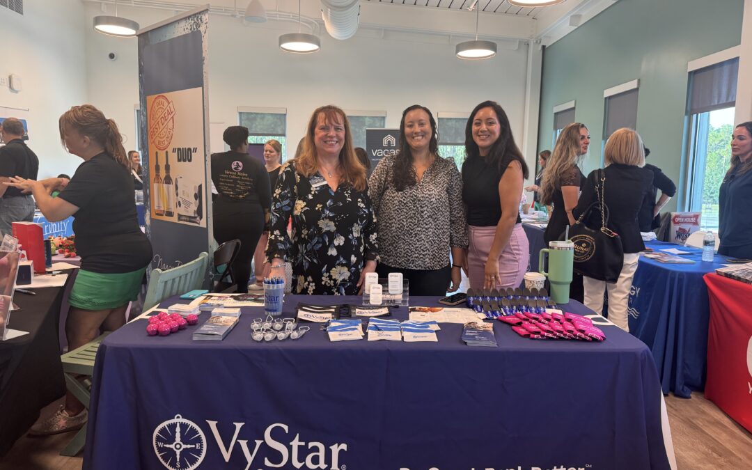 Three women stand behind a VyStar Credit Union table at a community event The table is covered with promotional items like pens, brochures, and keychains Other booths and people can be seen in the background The banner reads "Do Good Bank Better