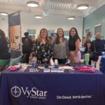 Three women stand behind a VyStar Credit Union table at a community event The table is covered with promotional items like pens, brochures, and keychains Other booths and people can be seen in the background The banner reads "Do Good Bank Better