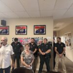 A group of people stands in a room with framed awards on the wall In the foreground, a man speaks into a microphone next to a woman in a gray shirt Behind them, police officers and men in casual wear stand facing forward A pink "the " banner is partially visible