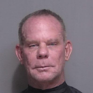 A close up photograph of a middle aged man with short, light hair He has a neutral facial expression and is wearing a dark colored shirt The background is a plain gray wall