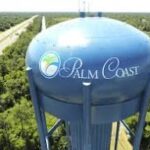 A tall blue water tower with "Palm Coast" and a green and blue logo on its side stands amidst a forested area with a road running through greenery in the background The sky is clear, suggesting a sunny day