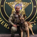 A sheriff's office member in tactical gear poses with a German Shepherd police dog in front of a sheriff's office emblem backdrop The man is holding the dog's leash, and both are looking at the camera The emblem reads "Sheriff's Office Flagler County