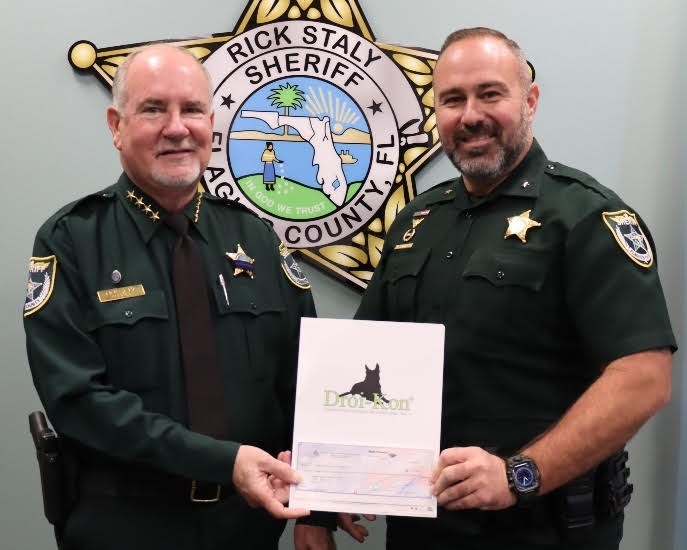 Two uniformed sheriffs stand in front of a sheriff department emblem The sheriff on the left holds a document with the "Droneten" logo and a signature line, while both smile at the camera