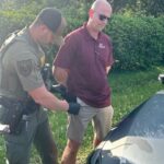 A law enforcement officer in a green uniform and black gloves is conducting a pat down search on a man in a maroon polo shirt and khaki shorts The man, standing by a vehicle and wearing sunglasses, has his hands behind his back Green foliage is visible in the background