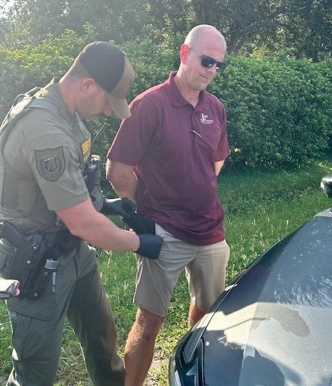 A law enforcement officer in a green uniform and black gloves is conducting a pat down search on a man in a maroon polo shirt and khaki shorts The man, standing by a vehicle and wearing sunglasses, has his hands behind his back Green foliage is visible in the background