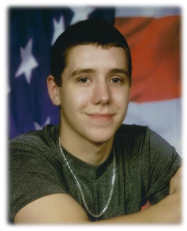 A young man with short dark hair, wearing a gray t shirt and a silver chain necklace, smiles while posing in front of an American flag backdrop