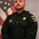 A uniformed officer stands in front of an American flag He has short dark hair, a trimmed beard, and mustache He wears a dark green uniform adorned with several pins and badges, including a gold star on his chest and shoulder patches He faces forward with a serious expression