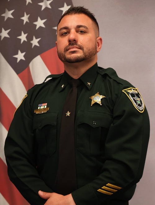 A uniformed officer stands in front of an American flag He has short dark hair, a trimmed beard, and mustache He wears a dark green uniform adorned with several pins and badges, including a gold star on his chest and shoulder patches He faces forward with a serious expression