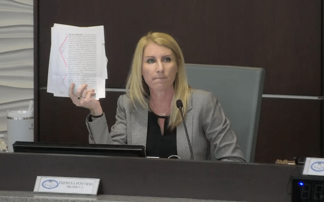 A person with long blonde hair is seated at a desk holding up a stack of papers in their left hand The individual is dressed in a grey blazer and a black top There are nameplates, microphones, and electronic devices on the desk in front of them