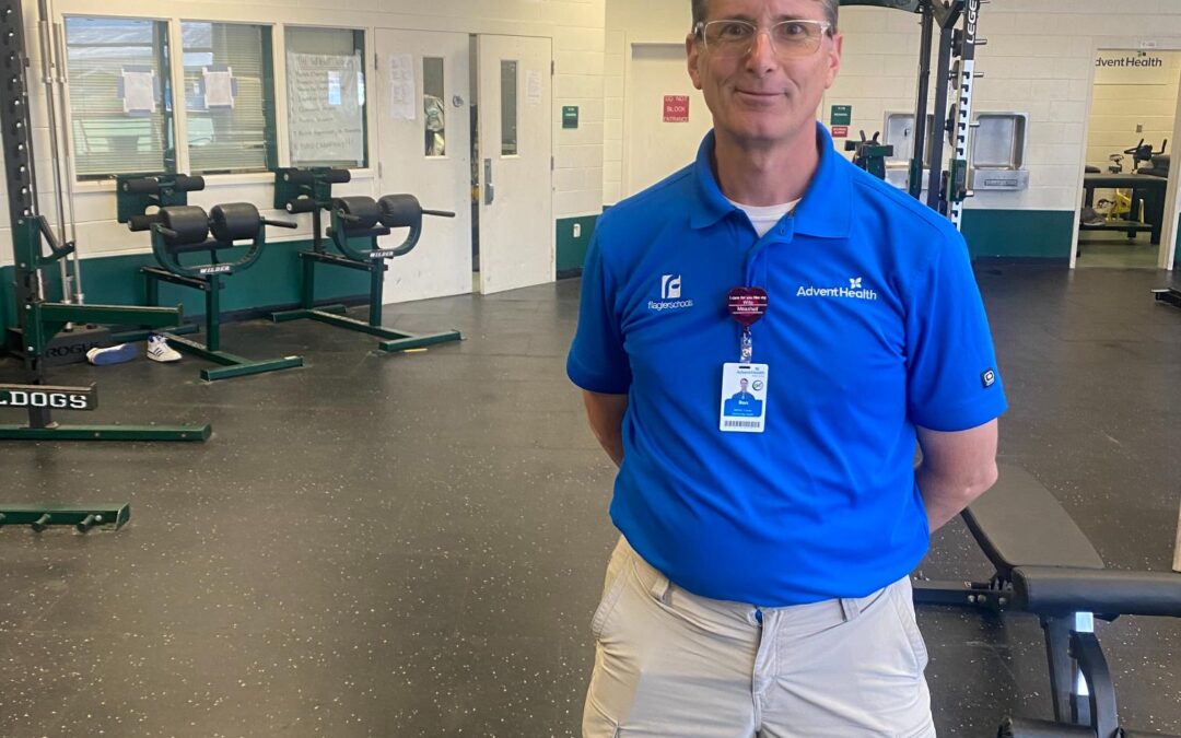 A man wearing a blue polo shirt and khaki shorts stands in a gym Behind him, the wall displays motivational words "BE GREAT" and the school's mascot name "BULLDOG " Gym equipment and weights are visible throughout the room