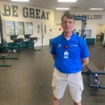 A man wearing a blue polo shirt and khaki shorts stands in a gym Behind him, the wall displays motivational words "BE GREAT" and the school's mascot name "BULLDOG " Gym equipment and weights are visible throughout the room