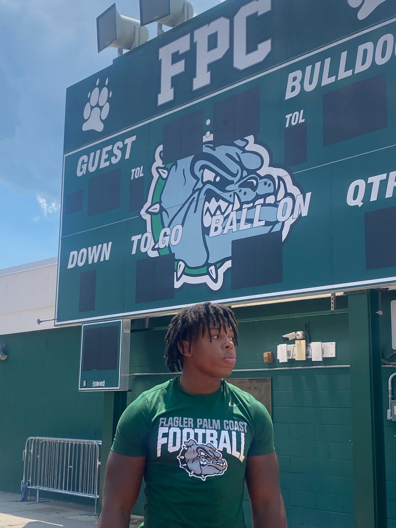 A young man wearing a green "Flagler Palm Coast Football" shirt stands in front of a large scoreboard The scoreboard features a bulldog mascot and sections labeled "Guest," "Home," "To Go," and "Ball On " The sky is clear and blue