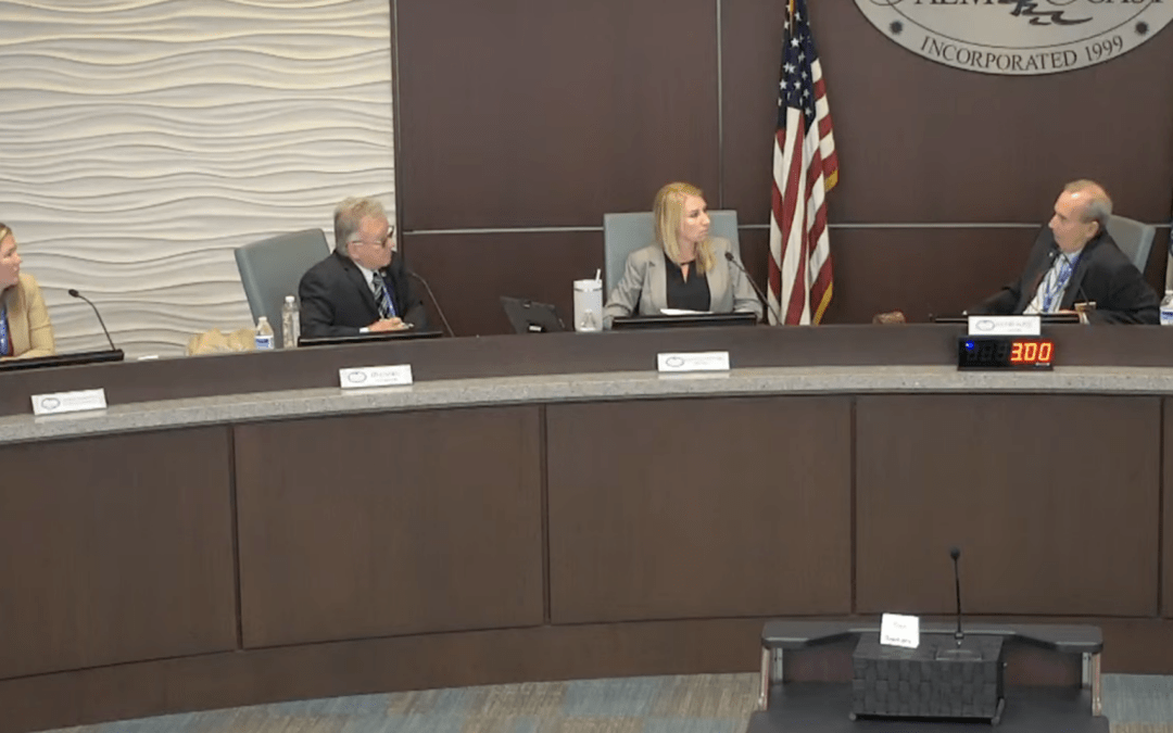 Four people are seated at a curved table in a government meeting room Three individuals are on one side, facing toward a person seated across from them Behind them is a wall with the American flag and a seal A digital clock displays ":