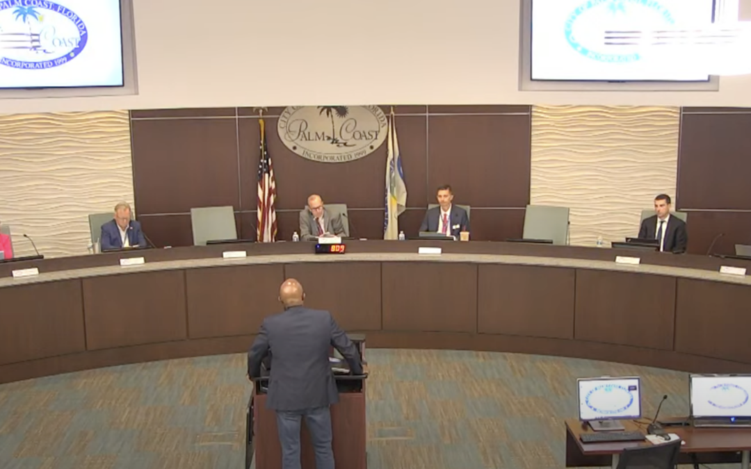 A man stands at a podium speaking to a panel of six seated individuals during a council meeting in Palm Coast The city logo is displayed on the wall behind the council members Several computer monitors and flags are visible in the background