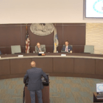 A man stands at a podium speaking to a panel of six seated individuals during a council meeting in Palm Coast The city logo is displayed on the wall behind the council members Several computer monitors and flags are visible in the background