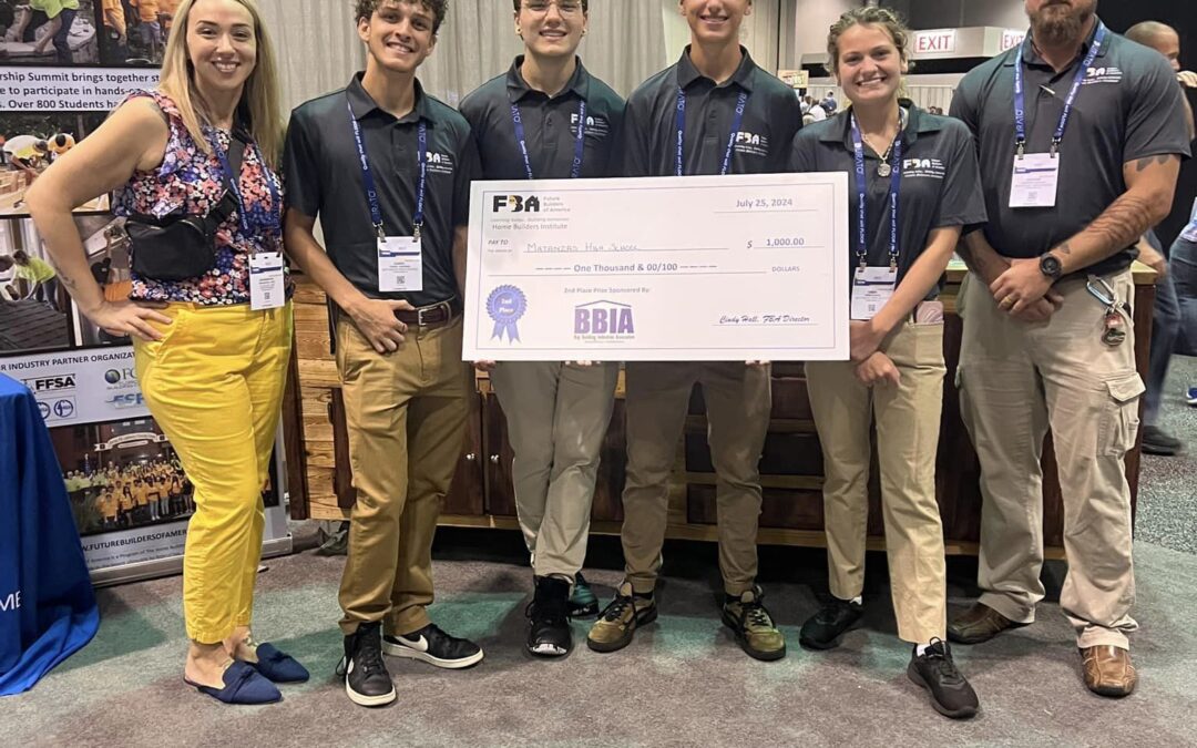 A group of five people stands together, holding a large check at an indoor event They are all smiling and wearing event badges Behind them is a booth displaying banners and informational material The environment appears to be a convention or conference setting