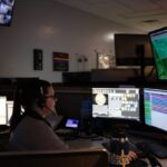 A person sits at a desk wearing a headset and is surrounded by multiple computer monitors displaying maps, data, and various graphical interfaces The room has dim lighting with additional screens mounted on the wall showing different perspectives