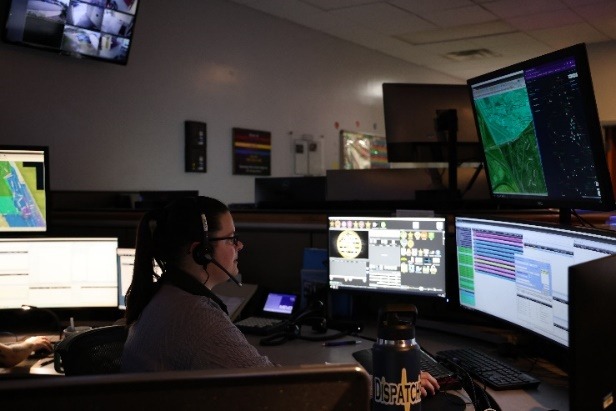 A person sits at a desk wearing a headset and is surrounded by multiple computer monitors displaying maps, data, and various graphical interfaces The room has dim lighting with additional screens mounted on the wall showing different perspectives