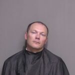 A man with short hair is shown against a plain background He is wearing a dark, draped garment, often associated with mugshots His expression is neutral, and he is facing the camera directly