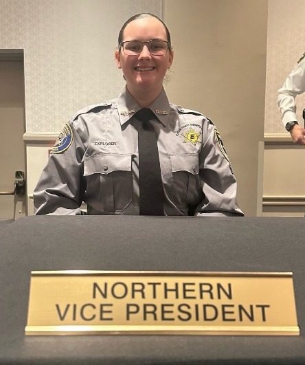 A person in a uniform, with a badge and patches, is smiling while seated behind a table with a nameplate that reads "NORTHERN VICE PRESIDENT " The person has glasses and their hair tied back The background includes a wall and a partially visible standing individual