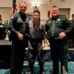 A young man stands proudly between two uniformed officers, wearing a medal around his neck The officers, dressed in dark green uniforms, smile beside him U S and state flags are visible in the background, with tables and people in the room