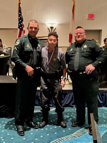 A young man stands proudly between two uniformed officers, wearing a medal around his neck The officers, dressed in dark green uniforms, smile beside him U S and state flags are visible in the background, with tables and people in the room