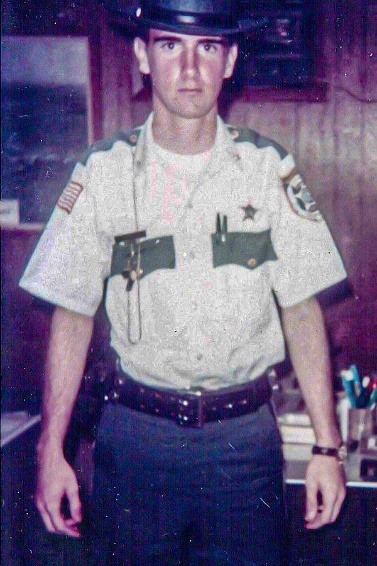 A man in a police uniform stands indoors, looking directly at the camera He wears a hat and a shirt with shoulder patches and a badge, along with a belt The background features a wooden wall with shelves containing various items