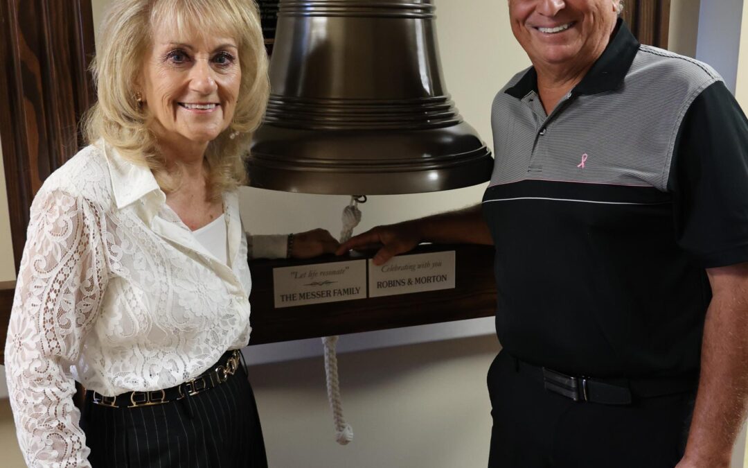 A woman and a man stand smiling in front of a large bell mounted on a wooden frame The man wears a dark polo shirt, and the woman wears a white blouse and striped pants