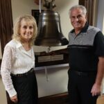 A woman and a man stand smiling in front of a large bell mounted on a wooden frame The man wears a dark polo shirt, and the woman wears a white blouse and striped pants