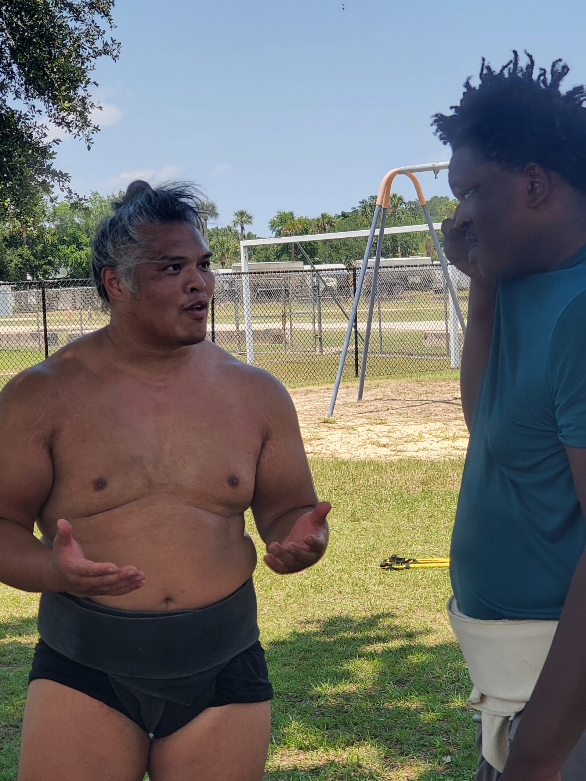 Two people stand outdoors on grass One is wearing a sumo outfit, engaging in conversation The other, dressed casually, listens A swing set and trees are in the background under a clear sky